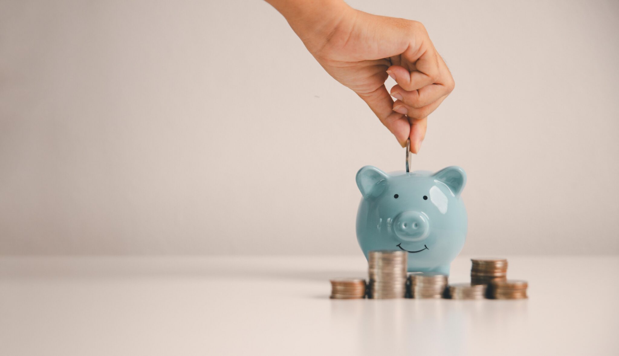 Hand putting a coin into a blue piggy bank next to coin stacks, illustrating personal finance and savings tips.